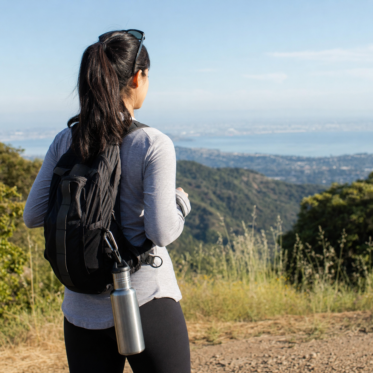 Hiker in gray activewear pauses at a mountain overlook, a metal water bottle clipped to their black daypack.