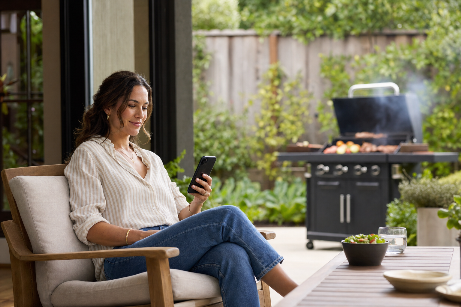 Woman relaxes in a patio chair checking her phone while food cooks unattended on a grill in the background